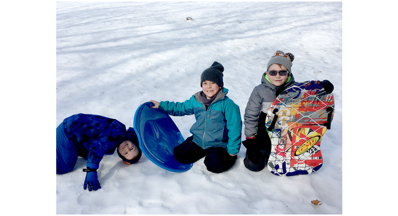 three smiling kids with sleds sitting in the snow